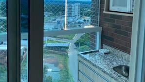 A kitchen with a microwave on a shelf, a window with a view of buildings, and a blue trash bin. - Condomínio Piazza diRoma Acqua Park Splash (Caldas Novas)