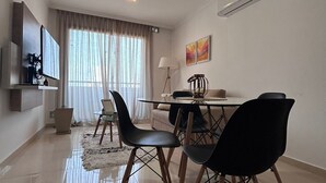 Una sala de estar moderna con comedor, que incluye un televisor de pantalla plana, una mesa de comedor con sillas y un ventanal con cortinas.