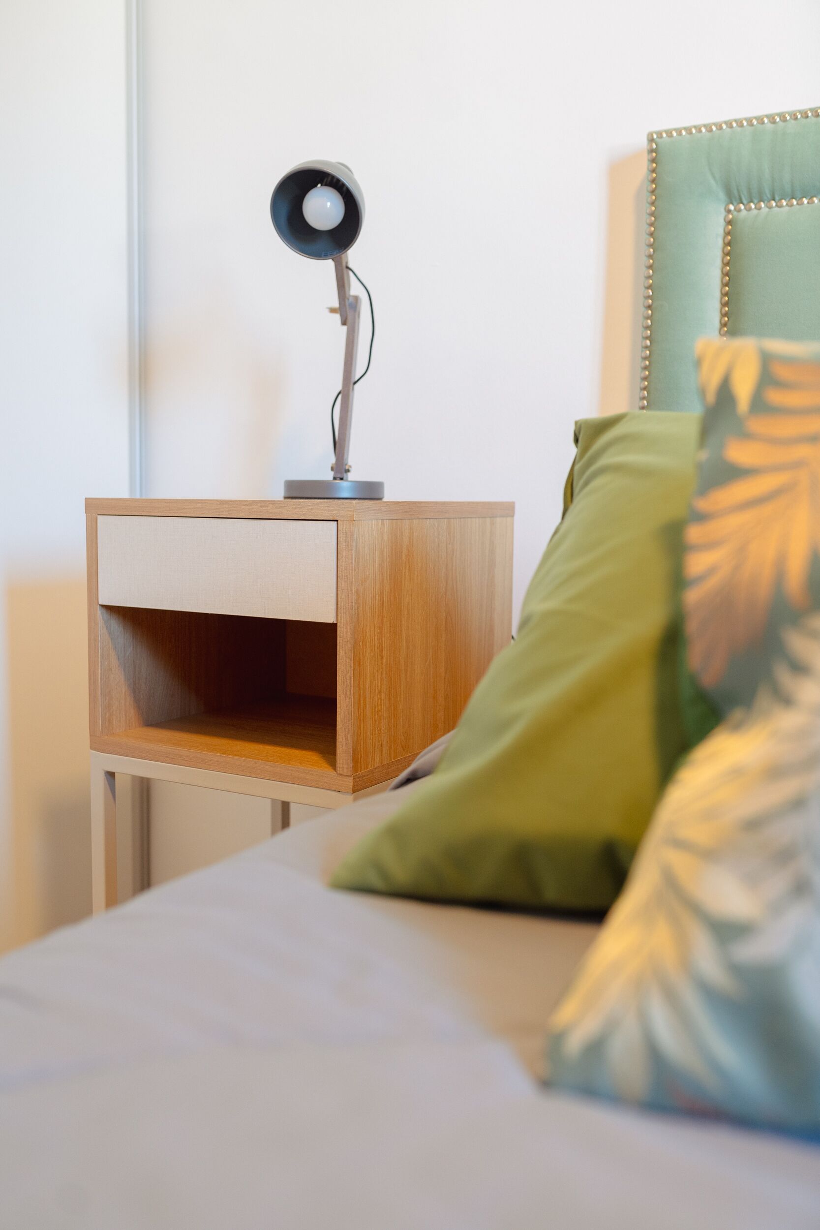 A neatly made bed with a green pillow, a bedside table with a lamp, and a wooden nightstand.