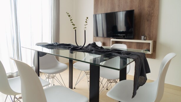 Un comedor moderno con mesa de vidrio, sillas blancas, un televisor de pantalla plana montado en un panel de madera y una planta en la esquina.