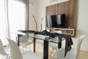 A modern dining area with a glass table, white chairs, a flat-screen TV mounted on a wooden panel, and a plant in the corner. - Agustín 1 Apart (Parana)