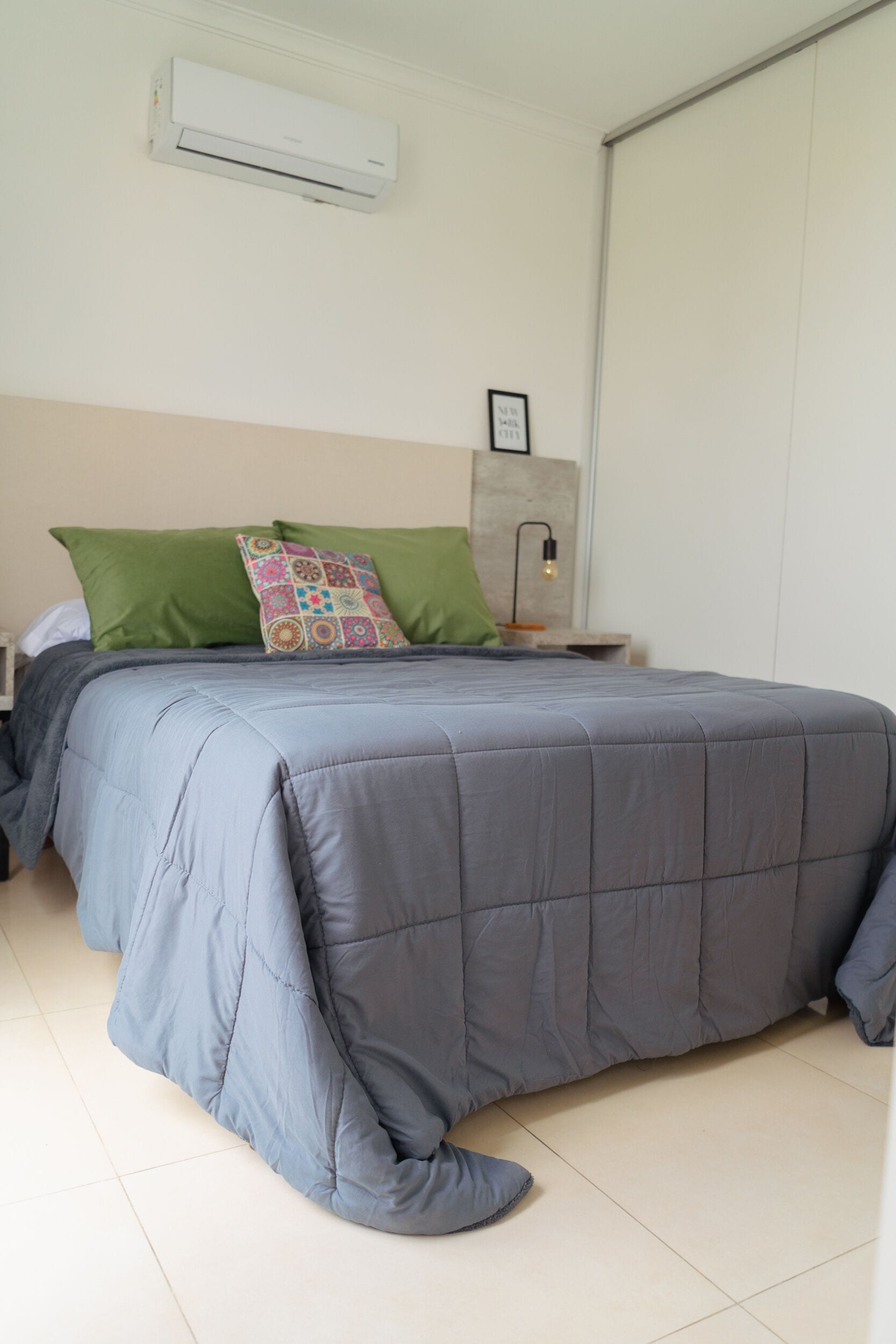 Une chambre à coucher avec un lit, des oreillers verts et multicolores, une lampe de chevet et un climatiseur fixé au mur.