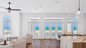A spacious room with a wooden floor, a dining area with a table and chairs, a sofa, and a view of the ocean through large glass doors.