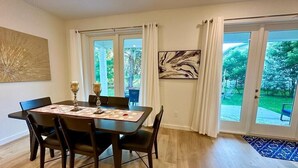 A dining area with a dark wooden table and chairs, two large windows with white curtains, and a view of a green outdoor area.