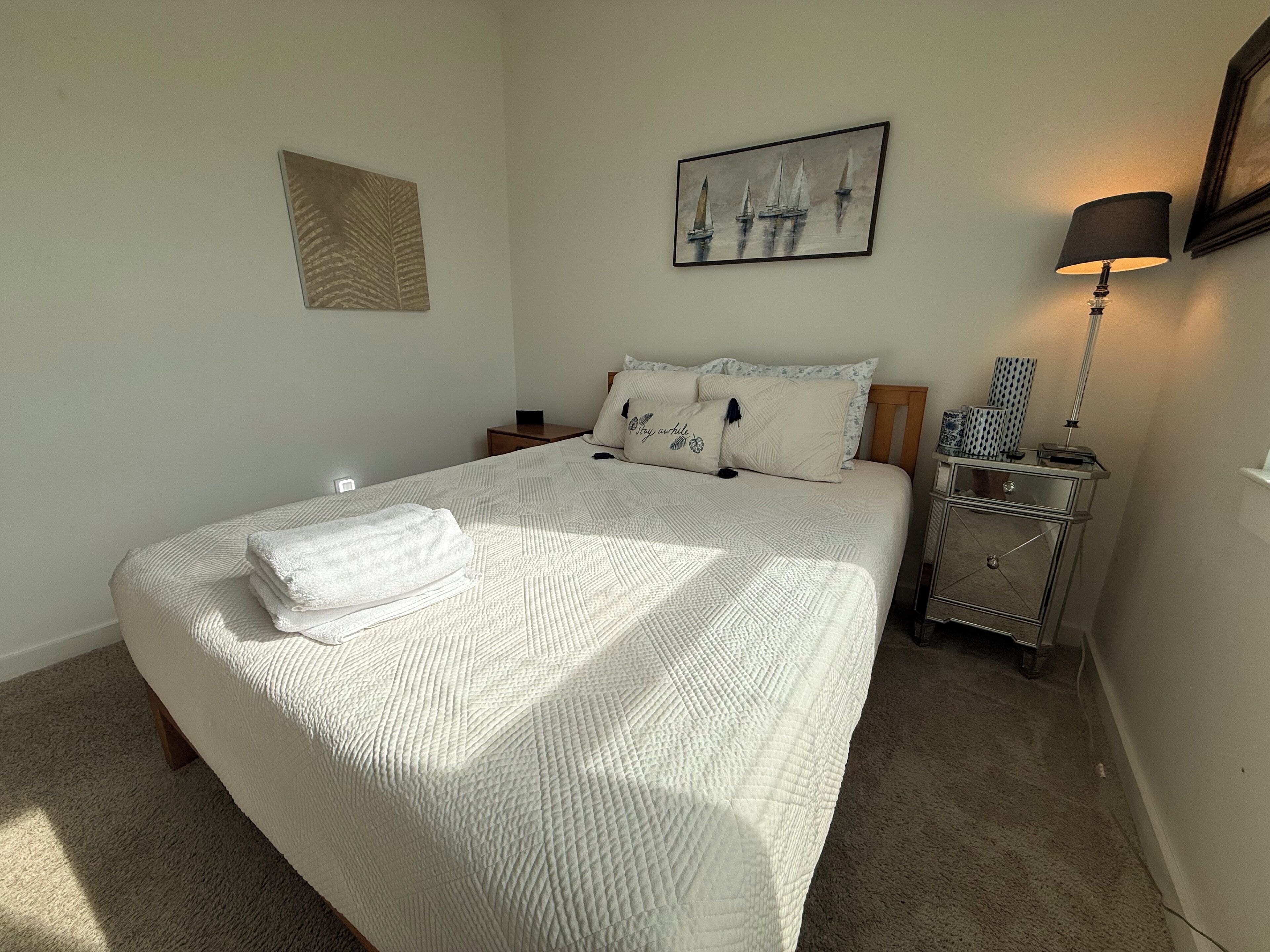Una cama bien hecha con colcha blanca, una mesita de noche con lámpara y una obra enmarcada sobre la cama.