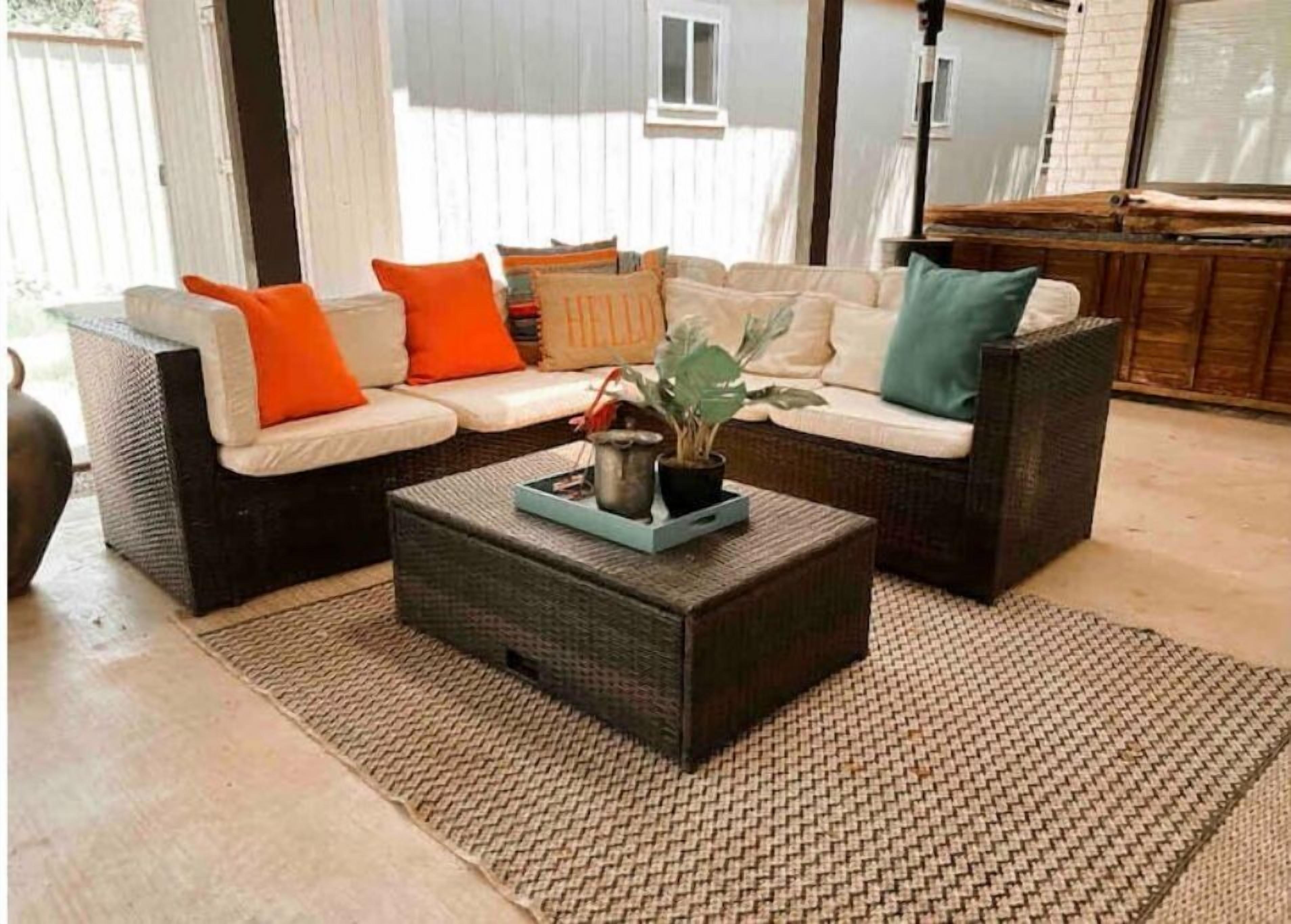 Ein Ecksofa mit orangefarbenen und türkisfarbenen Kissen, ein dunkler Korbtisch mit Tablett und ein gewebter Teppich.