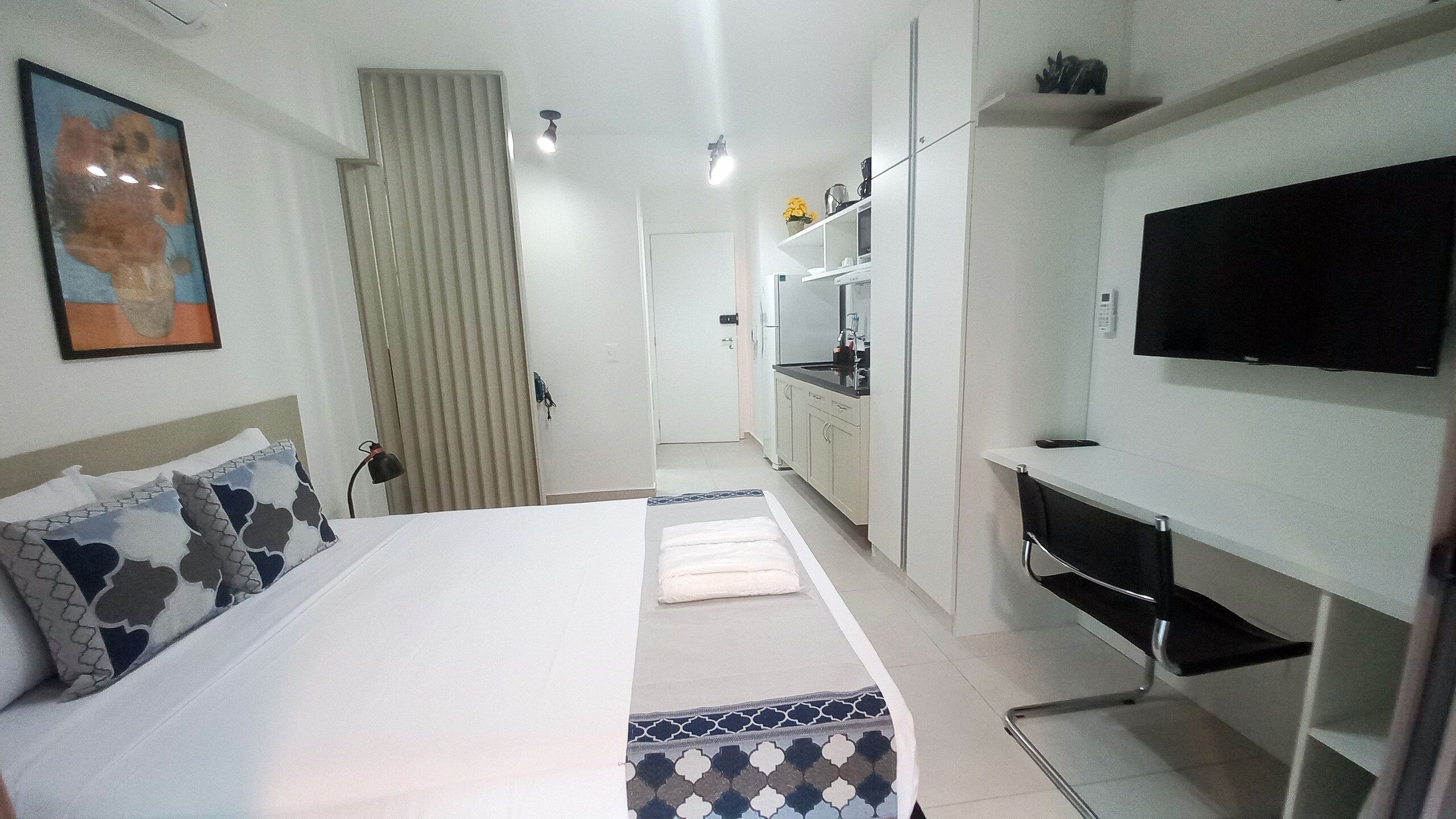 A modern hotel room with a bed, a desk, a TV, and a kitchenette.