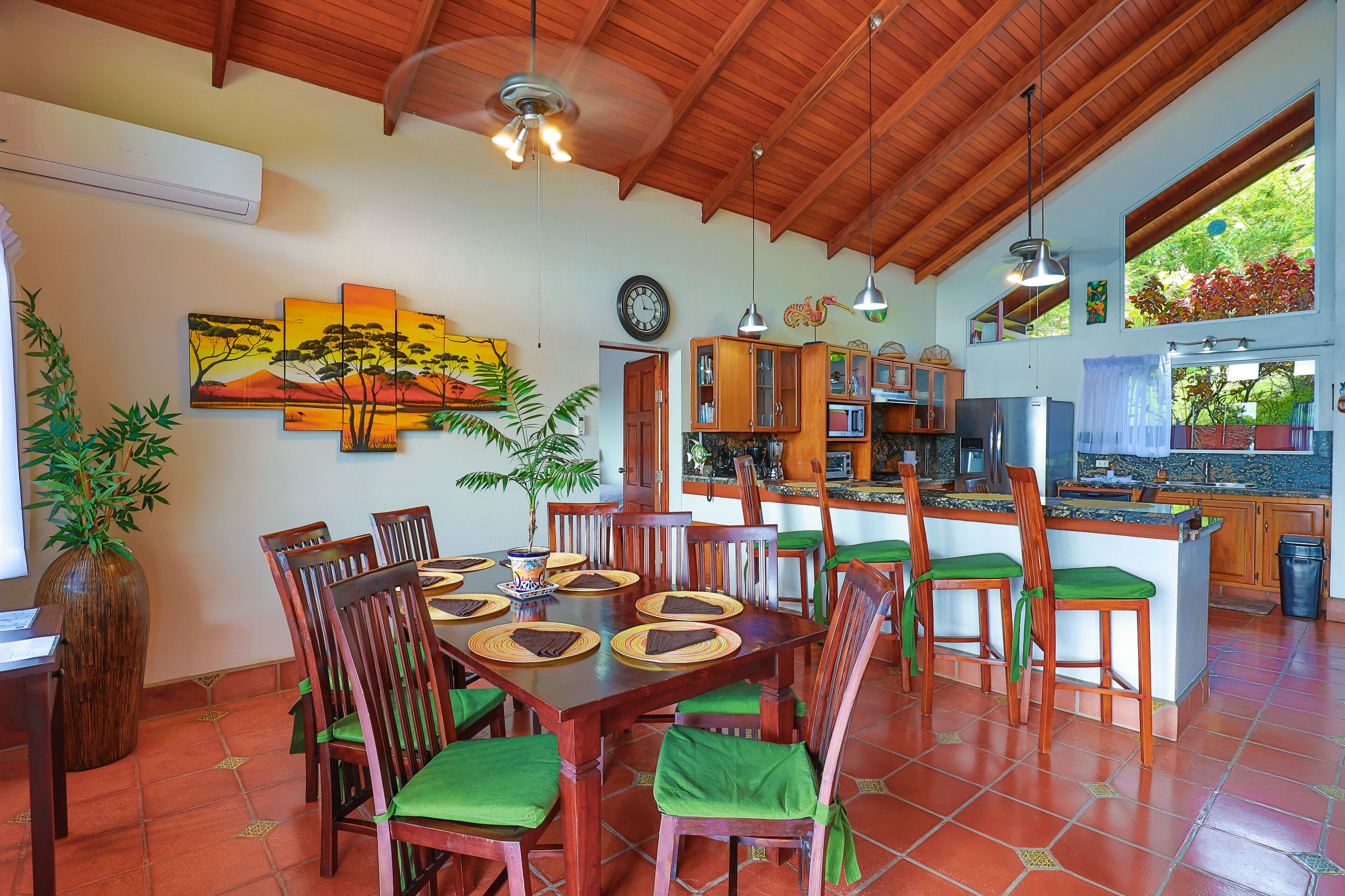 A spacious dining area with a wooden table and chairs, a large painting on the wall, and a kitchen in the background.
