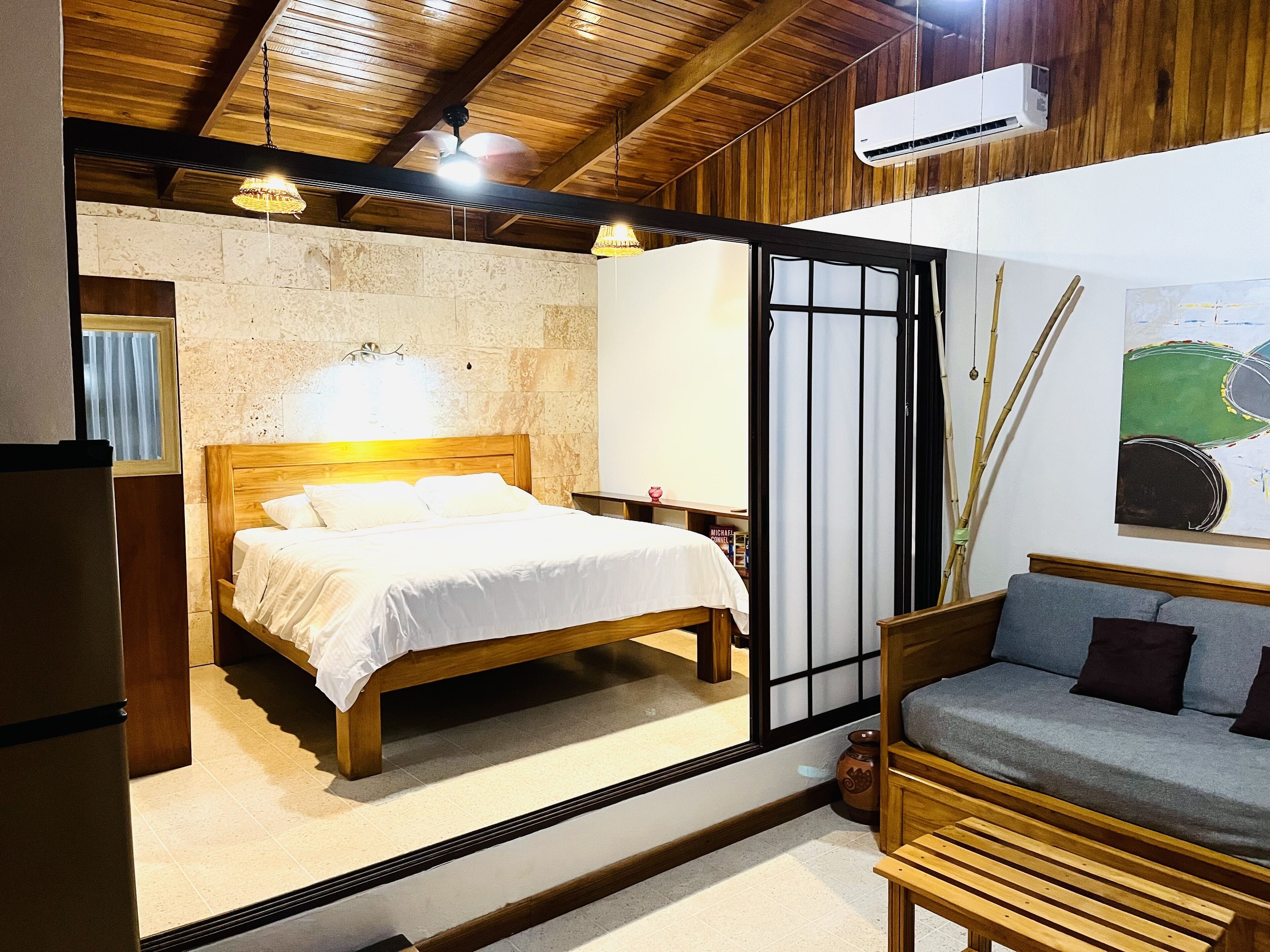 A bedroom with a wooden bed, a sofa, a stone wall, and a large window.