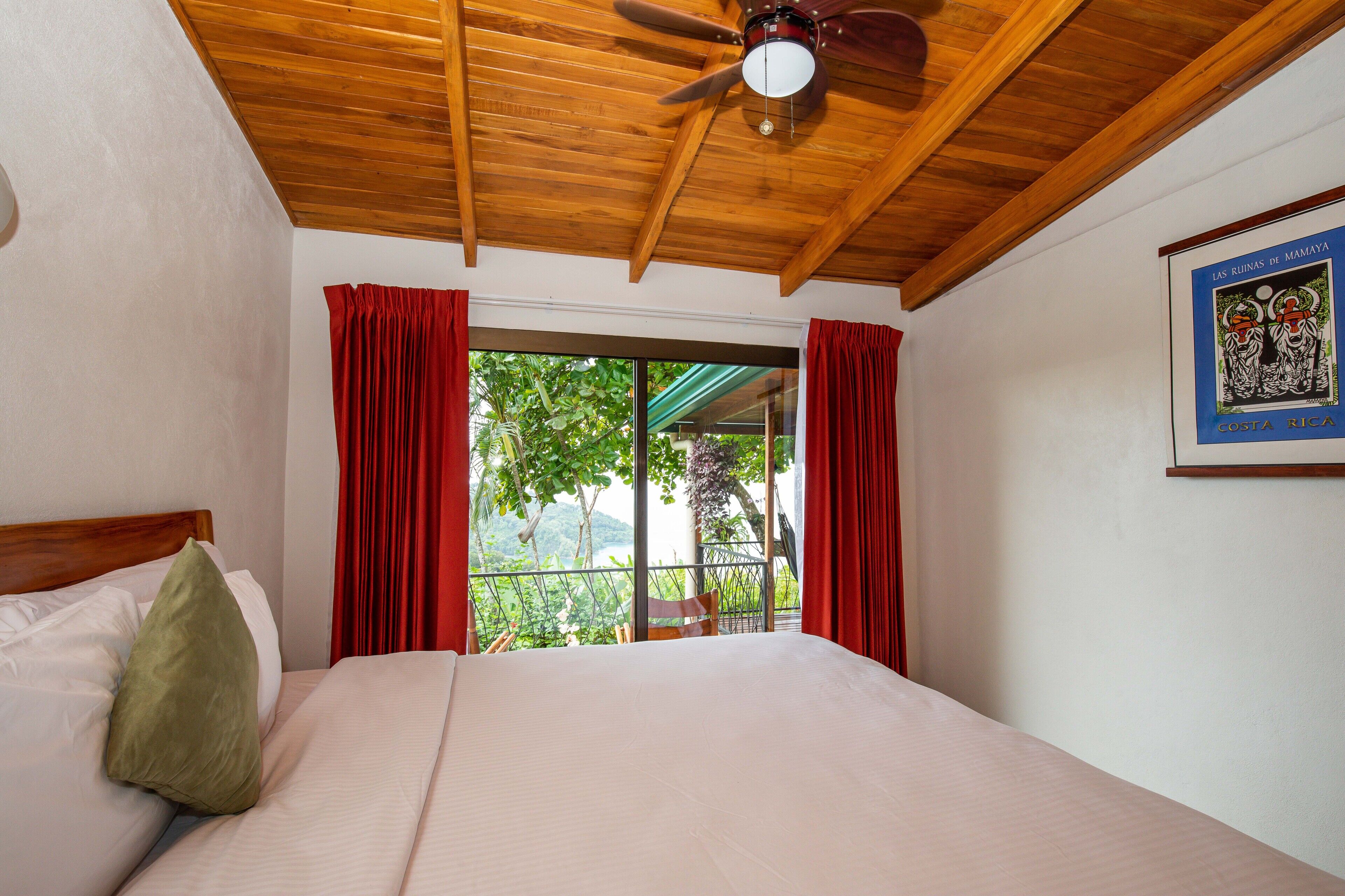 A hotel room with a wooden ceiling, a bed with white linens, red curtains, and a view of greenery outside.