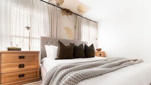 Une chambre à coucher avec un lit, une table de chevet en bois, une lampe et une fenêtre avec des rideaux.
