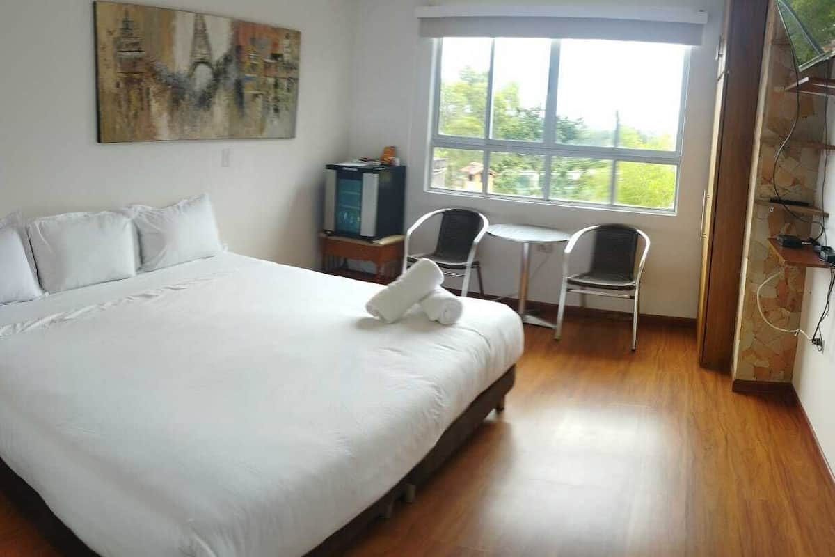 A hotel room with a large bed, a small table, two chairs, a painting on the wall, and a window with a view of greenery.