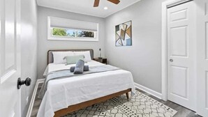 A bedroom with a bed, a ceiling fan, a window, a door, and a geometric artwork on the wall. - Relaxing Lake Zurich 3BR + Patio | LCP Collection (Lake Zurich)