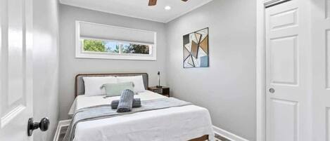 Un dormitorio con cama, ventilador de techo, ventana, puerta y una obra de arte geométrica en la pared.