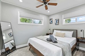 A bedroom with a bed, a ceiling fan, two bedside tables, a mirror, and windows with blinds. - Relaxing Lake Zurich 3BR + Patio | LCP Collection (Lake Zurich)