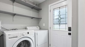 A laundry room with a front-loading washing machine and dryer, metal shelves above, and a door with a decorative glass window. - Relaxing Lake Zurich 3BR + Patio | LCP Collection (Lake Zurich)