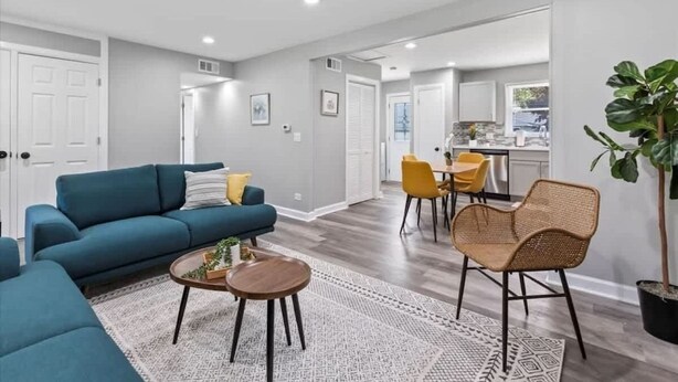 A modern living room with a blue sofa, a round coffee table, and a wicker chair. There is a kitchen in the background with yellow chairs. - Relaxing Lake Zurich 3BR + Patio | LCP Collection (Lake Zurich)