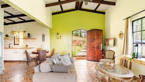 A spacious living area with a sofa, coffee table, and chairs. A kitchen area is visible in the background with a dining table and chairs.