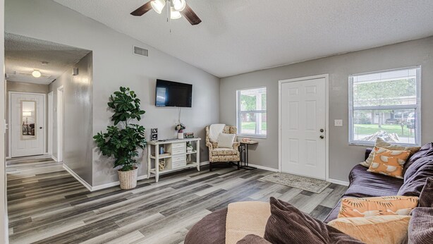 A spacious living room with hardwood flooring, a ceiling fan, a television, a comfortable sofa, and a large window with blinds.