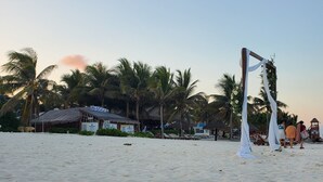 A beach with a wooden archway decorated with white fabric and flowers, palm trees, and a few buildings in the background.
