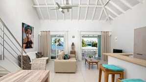 Un vaste espace de vie comprenant un canapé, une table basse et une table à manger. Un escalier se trouve sur la gauche, ainsi qu’un balcon offrant une vue sur la mer et un ventilateur de plafond.