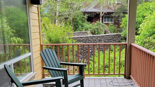 Eine Holzterrasse mit zwei Adirondack-Stühlen und einem kleinen Tisch, mit Blick auf eine Steinmauer und üppiges Grün.