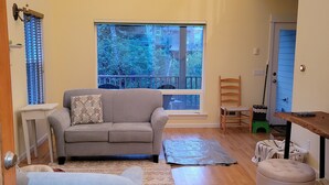 A spacious living room with a grey sofa, a blue armchair, and a large window offering a view of trees. - Yachats Beach Cottage -- 5 min walk to the ocean. (Yachats)