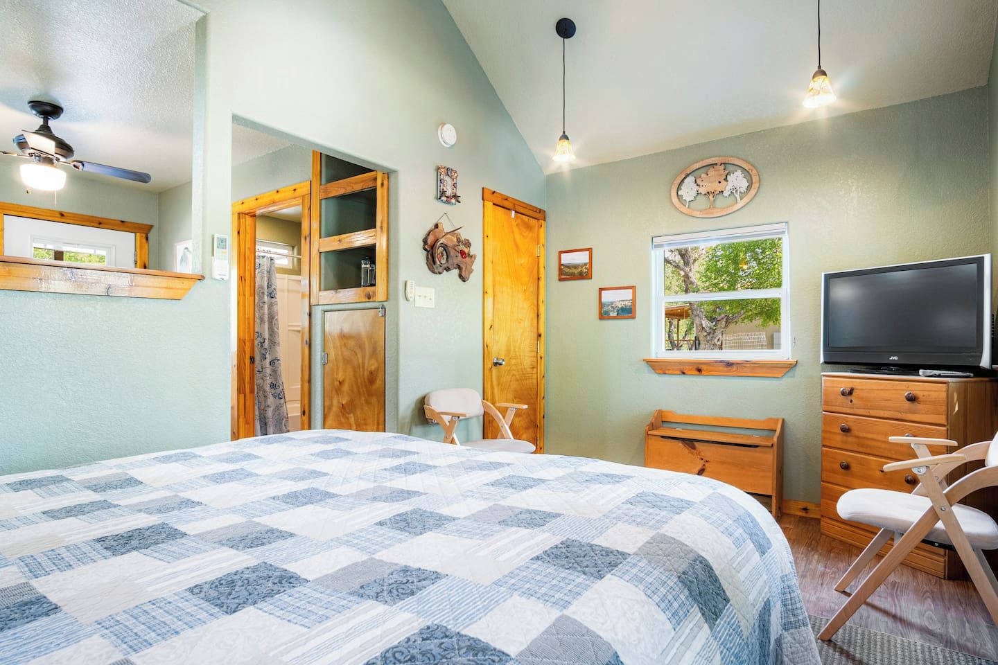 A bedroom with a large bed, a television, a wooden dresser, and a window with a view of trees.