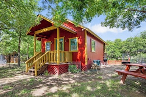 Una cabaña roja con un porche de madera y una escalera, rodeada de árboles y una mesa de picnic.