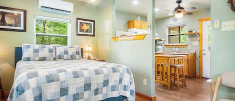 A bedroom with a bed, bedside lamps, a window with a view, and a kitchenette area with a ceiling fan.