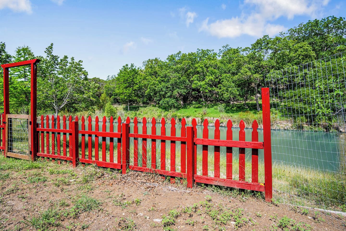 A red metal fence with a gate, surrounded by greenery and a body of water.