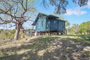 A two-story house with a balcony, surrounded by trees and grassy terrain. - Stronghold Crazy Horse | Cabin w/ Hot Tub & Creek (Bandera)