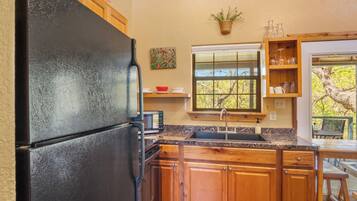 A kitchen with wooden cabinets, a black refrigerator, a microwave, a sink, and a window with a view of trees.