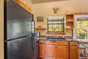 A kitchen with wooden cabinets, a black refrigerator, a microwave, a sink, and a window with a view of trees. - Stronghold Crazy Horse | Cabin w/ Hot Tub & Creek (Bandera)