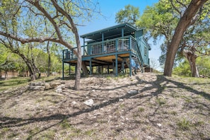 A two-story building with a balcony, surrounded by trees and a rocky ground.