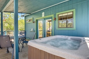 A hot tub under a covered patio with a table and chairs, wooden deck, and a door with a sign. - Stronghold Crazy Horse | Cabin w/ Hot Tub & Creek (Bandera)