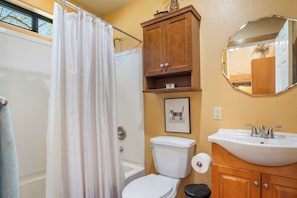 A bathroom with a shower curtain, toilet, cabinet, sink, and a framed picture on the wall. - Stronghold Crazy Horse | Cabin w/ Hot Tub & Creek (Bandera)