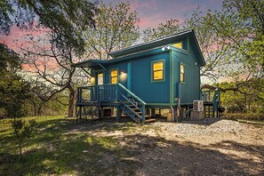 A two-story house with a porch and stairs, surrounded by trees and a gravel driveway. - Stronghold Crazy Horse | Cabin w/ Hot Tub & Creek (Bandera)