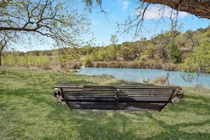 A bench in a grassy area with a view of a body of water and trees. - Stronghold Crazy Horse | Cabin w/ Hot Tub & Creek (Bandera)