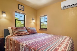 Un dormitorio con una cama, dos ventanas, aire acondicionado y una mesita de noche.