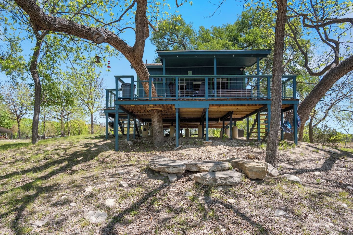 Ein Baumhaus mit einer Holzterrasse und einem Metallgeländer, umgeben von Bäumen und einem wolkenlosen Himmel.