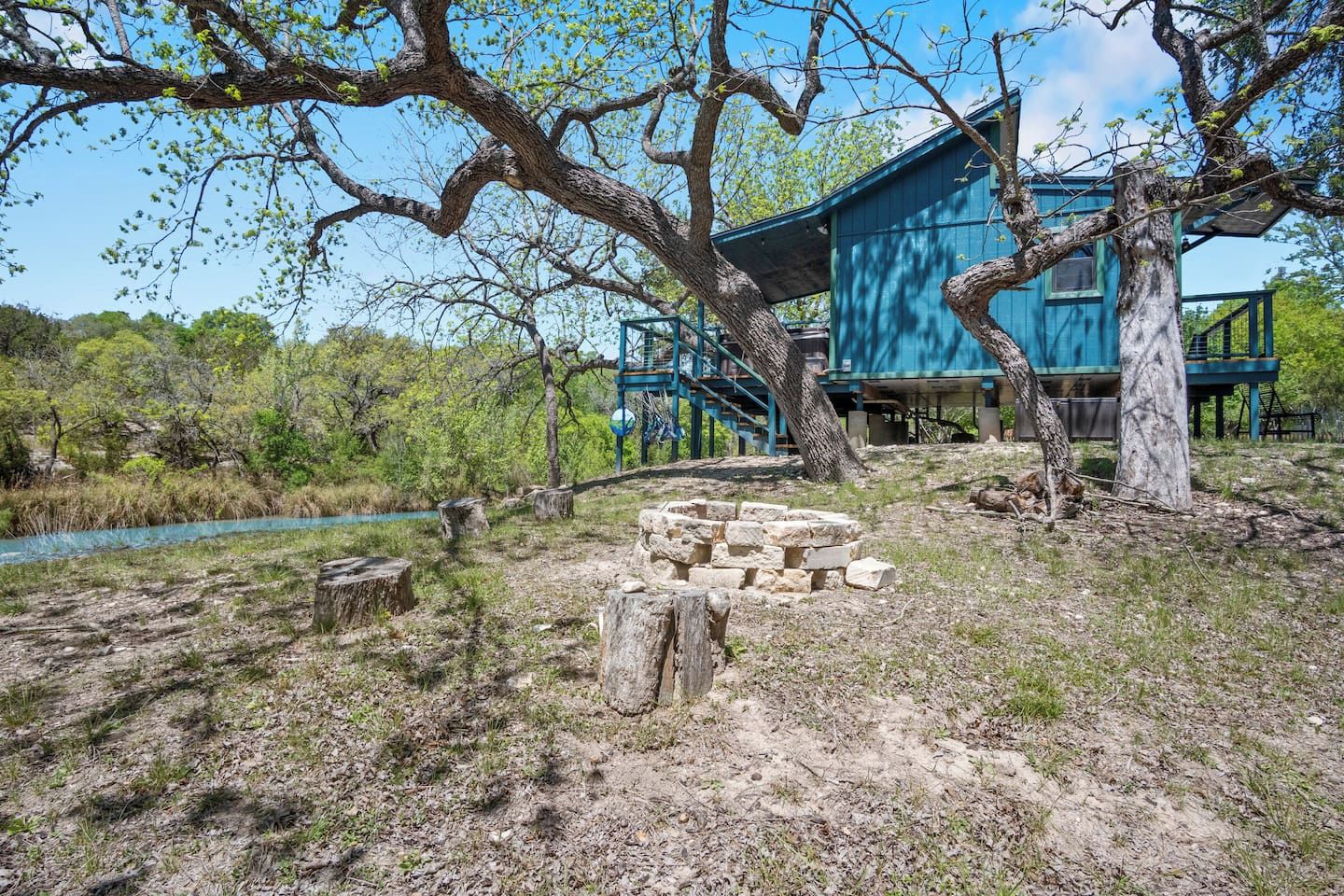A rustic outdoor area with a stone fire pit, a blue building with a staircase, and a natural landscape with trees and a water body.