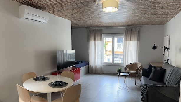 A modern living room with a sofa, a dining table with chairs, a TV, and a window with curtains. - Spacieux Studio Pour Toute la Famille (Palavas-les-Flots)
