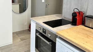 A modern kitchen with white cabinets, a built-in oven, a dishwasher, a black induction cooktop, and a red kettle. - Spacieux Studio Pour Toute la Famille (Palavas-les-Flots)