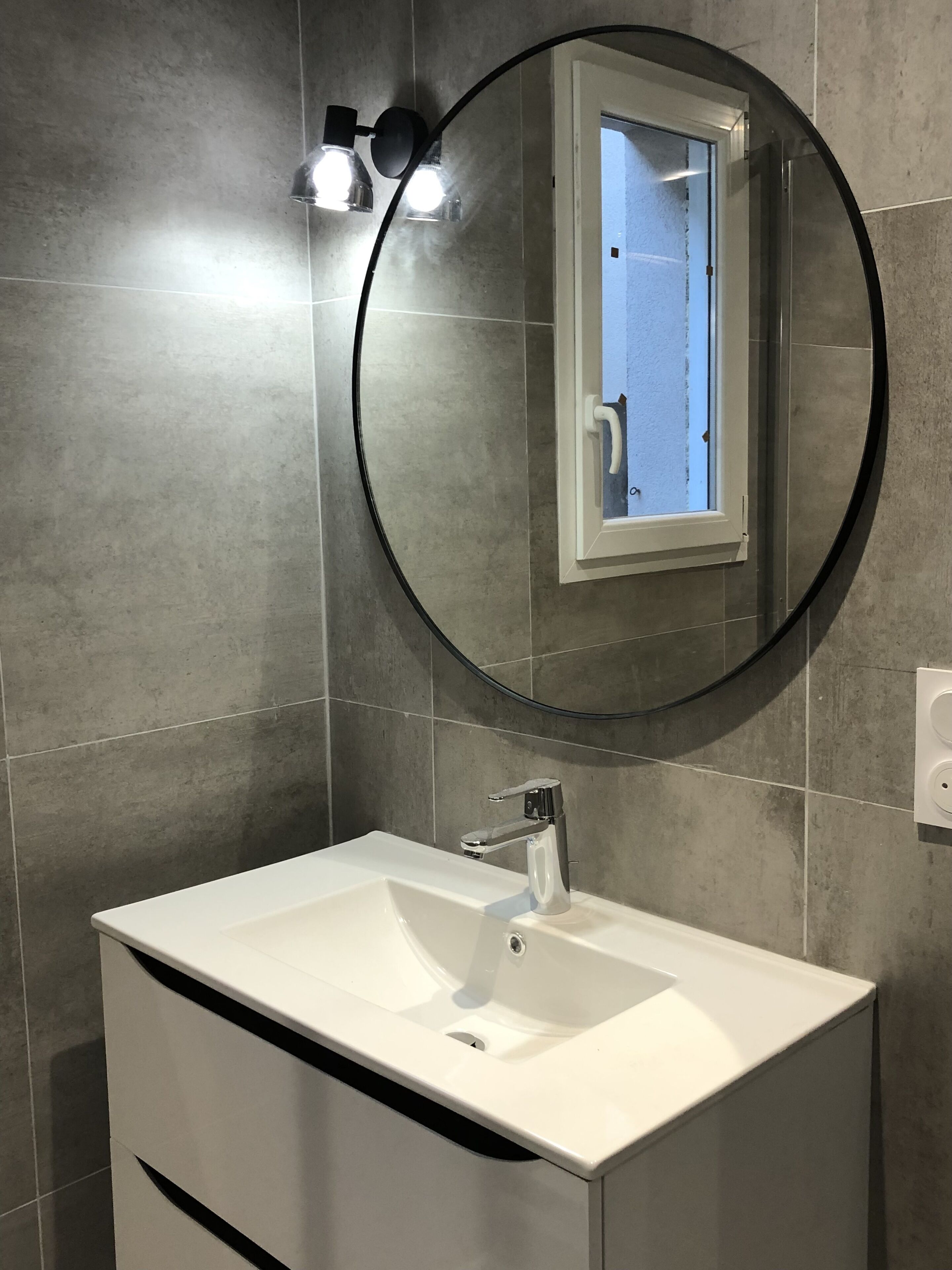 Une salle de bain avec un lavabo blanc, un miroir rond et un luminaire placé au-dessus du miroir.