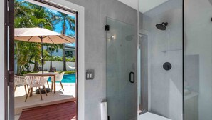 A modern bathroom with a glass-enclosed shower, a white sink, and a glass door leading to a pool area with outdoor furniture. - Surfside Oasis Pool home/5 min to Beach (Delray Beach)