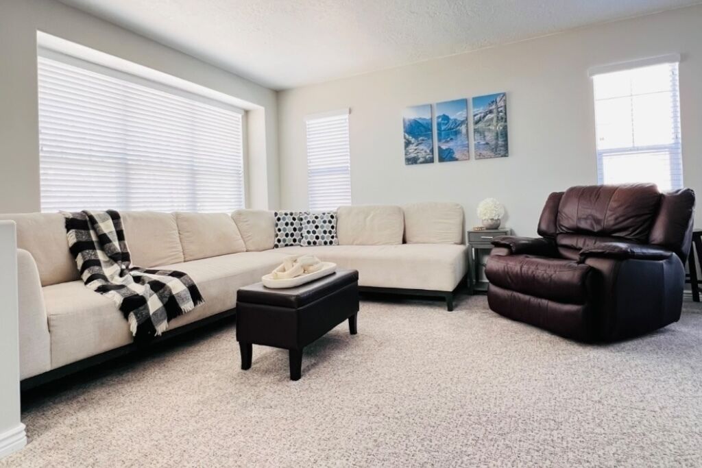 Een woonkamer met een beige bank, een bruine leren fauteuil, een klein tafeltje en een salontafel.