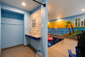 A bathroom with a blue wall, a wooden accent wall, a tiled sink, a shower area, and a mural of a sunset over the ocean. - Loveland Oasis w/ Indoor Heated Pool & Game Room (Loveland)