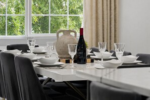 Mesa de comedor con platos, tazones y vasos blancos, una botella de vino Cato, y un florero texturizado.