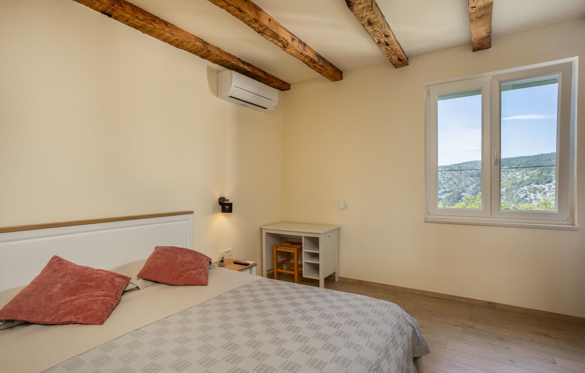 Quarto com cama, duas almofadas, mesa de cabeceira, pequena mesa, ar condicionado, janela com vista e vigas de madeira no teto.